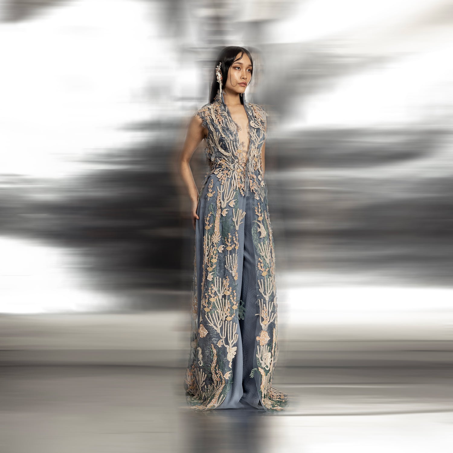 full length of Women's sleeveless reef-embroidered high neck jacket with floor-length panels over blue trousers – couture ensemble