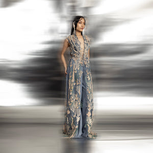 full length of Women's sleeveless reef-embroidered high neck jacket with floor-length panels over blue trousers – couture ensemble