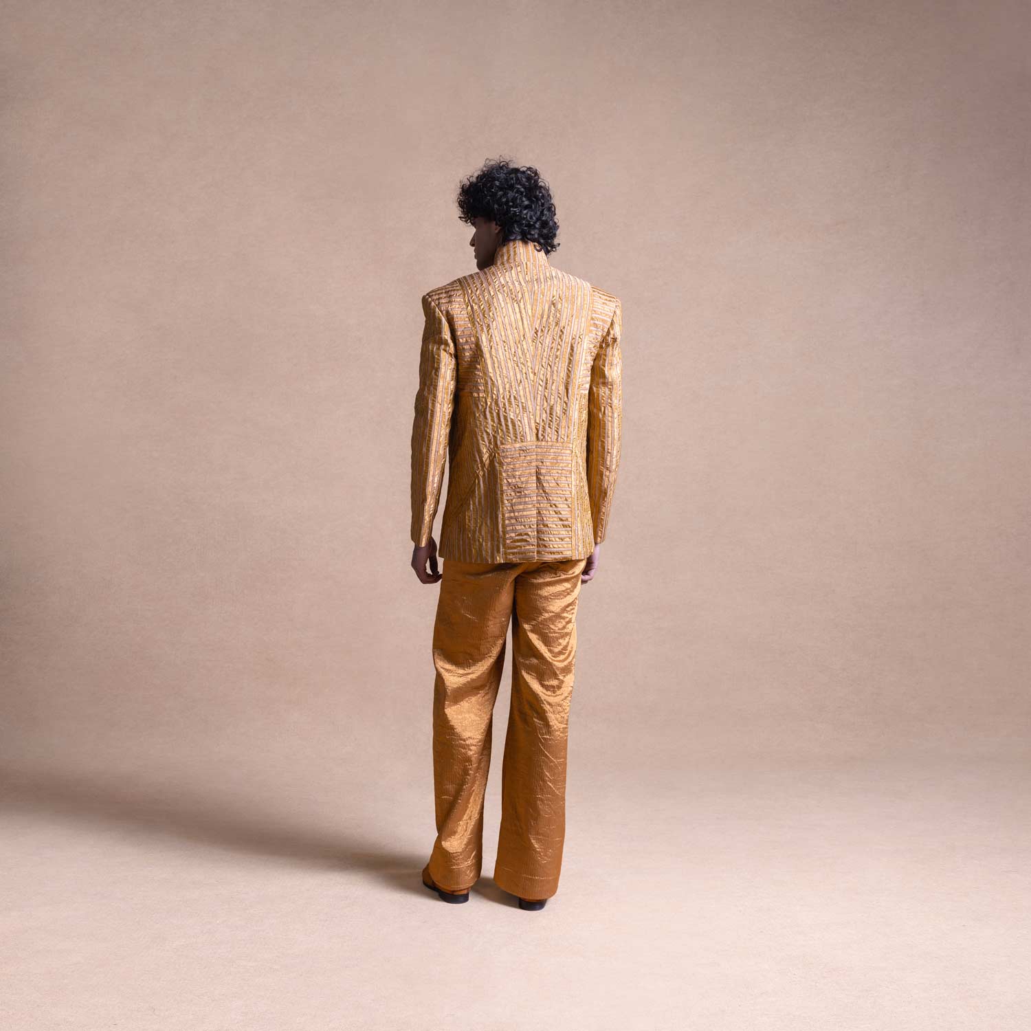 Back view of model wearing a gold textured Bandhgala suit with structured shoulders and wide-leg pants against a neutral beige backdrop - Tuxedo, retro suit, menswear
