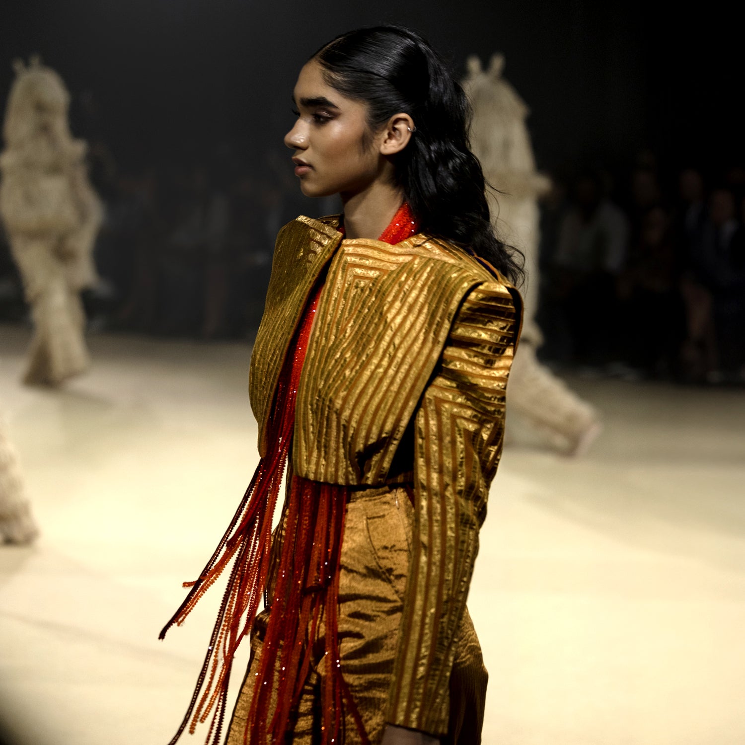Female model walking runway in a structured metallic gold jacket with exaggerated shoulders and flared pants - high fashion ensemble, powershoulder jacket, metallic.