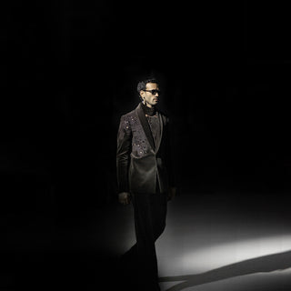 Men’s black cutwork tuxedo jacket with embellished lapel and tailored trousers under runway spotlight