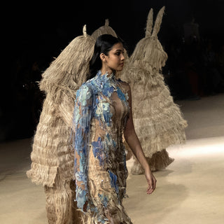 Model in floral-embellished nature-inspired gown walking past straw-costumed figures on runway - eco-luxury fashion, reception gown