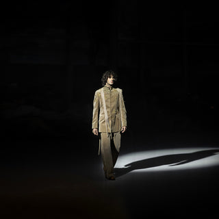 Person wearing a beige coat and pants walking on a dark runway with spotlight