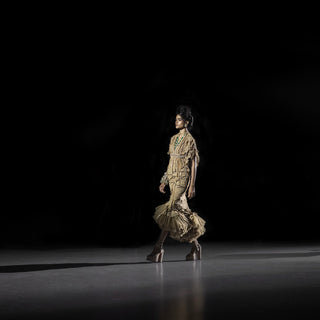 Women’s gold embellished cape and ruffled skirt set with layered detailing on runway under spotlight