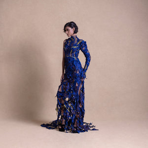 Model stands in a striking sheer blue gown with intricate textured appliqué and one long sleeve, - red carpet couture gown, cocktail gown, sheer embroidered gown