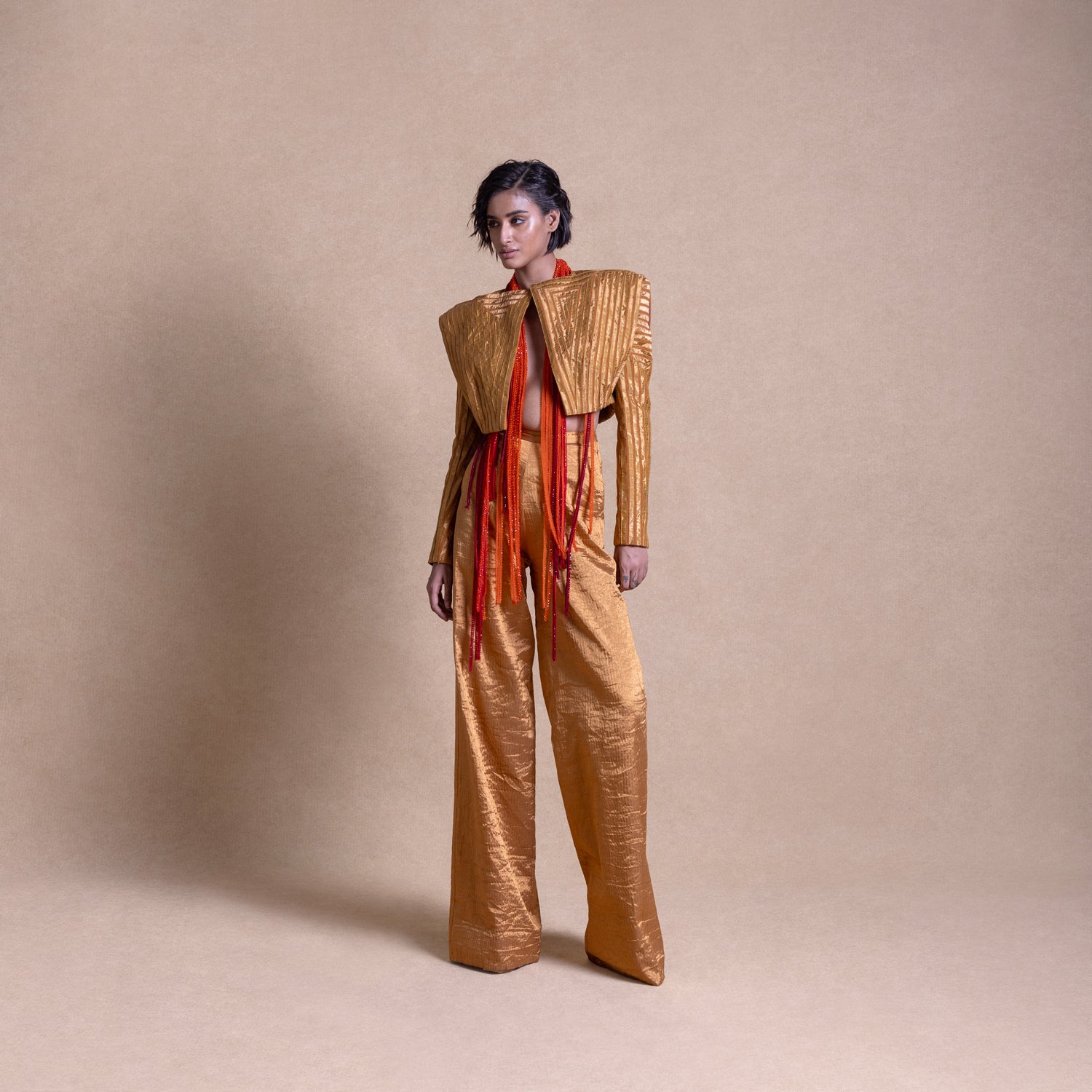 Model in gold structured jacket with exaggerated shoulders and matching wide-leg pants, adorned with red beaded fringe, standing against a beige studio backdrop - high fashion ensemble, powershoulder jacket, metallic