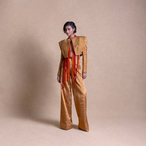 Model in gold structured jacket with exaggerated shoulders and matching wide-leg pants, adorned with red beaded fringe, standing against a beige studio backdrop - high fashion ensemble, powershoulder jacket, metallic