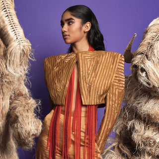 Model in Structured gold jacket with red beaded fringe details, posing between abstract straw creatures against a purple background - high fashion ensemble, powershoulder jacket, metallic.