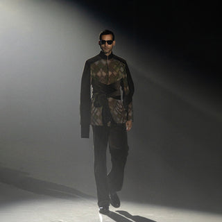 Male model in black patterned fashion jacket with draped detail and relaxed trousers on runway catwalk in spotlight