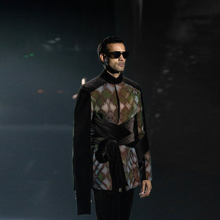 Man wearing a patterned shirt and dark coat with sunglasses against a dark background