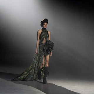 Women’s green patterned flowing evening dress with high slit and black boots walking runway under dramatic spotlight
