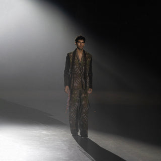 Men’s green patterned tailored suit with matching trousers walking runway under dramatic spotlight at fashion show